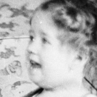Young child holding umbrella indoors, dressed in diaper and overalls.