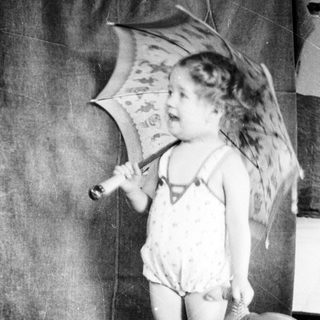 Young child holding umbrella indoors, dressed in diaper and overalls.