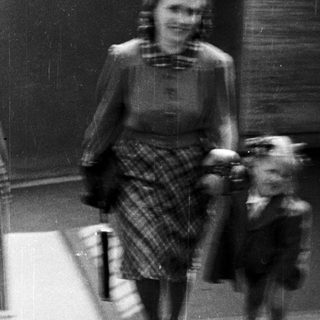 A woman in 1940s attire walks a small dog outdoors.