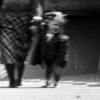 A woman in 1940s attire walks a small dog outdoors.