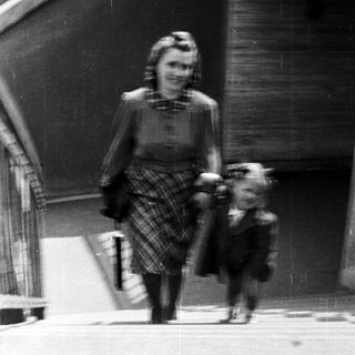 A woman in 1940s attire walks a small dog outdoors.