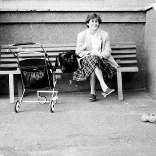 A woman sits on a bench with a stroller, a child holds a small dog on a leash.