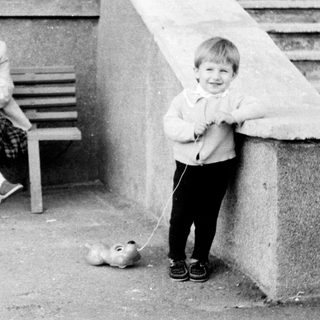 A woman sits on a bench with a stroller, a child holds a small dog on a leash.