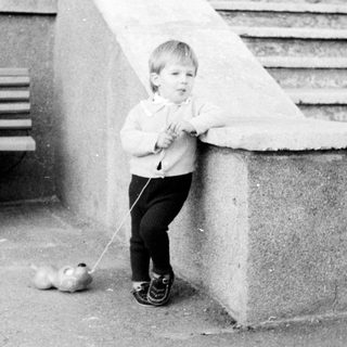 A child walks a small dog while an adult sits nearby.