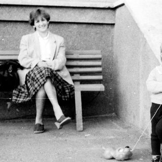 A child walks a small dog while an adult sits nearby.