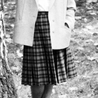 Woman standing outdoors near tree in 1960s-style attire.