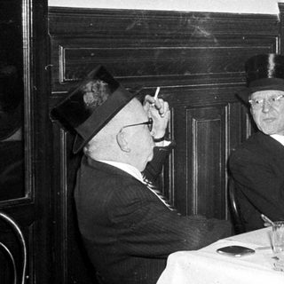 Two elderly men in formal attire smoking indoors.