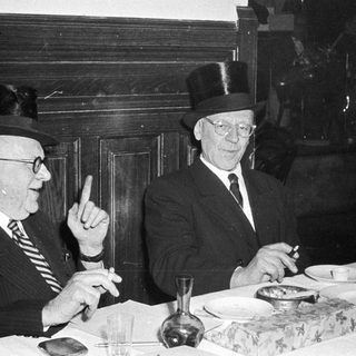 Two elderly men in formal attire having a meal together indoors.