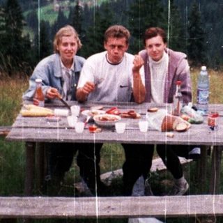 Three people picnic in rain on mountain meadow.
