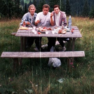 Three people picnic in rain on mountain meadow.
