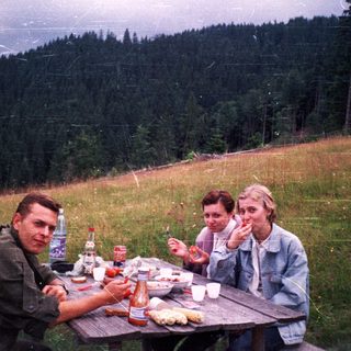 Three people picnicking outdoors on grassy hillside.