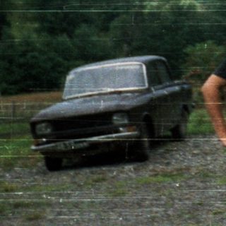 Three people posing outdoors near old car.