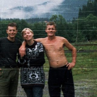 Three people posing outdoors near old car.