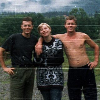 Three people posing outdoors near old car.