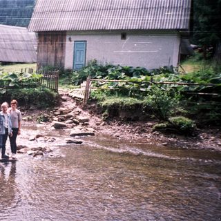 Two people standing by a shallow, rocky stream near rural houses.