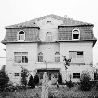 Two-story mansion with mansard roof, surrounded by fence.