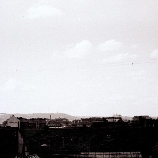 Black and white cityscape with visible rooftops and cloudy sky.
