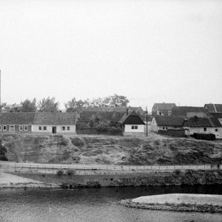 Black and white photo depicts village with tower overlooking water.