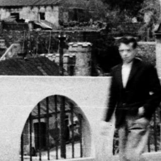 Black and white photo shows rooftops and a man by a bridge.