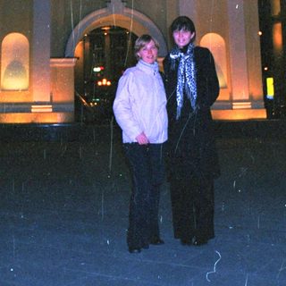 Two people stand in the rain near a building.