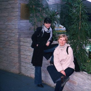 Two women pose near a fountain and stone wall.