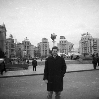 Man stands in a plaza with buildings and people.