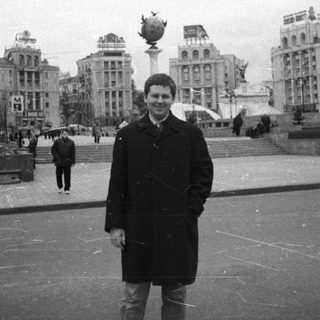 Man stands in a plaza with buildings and people.