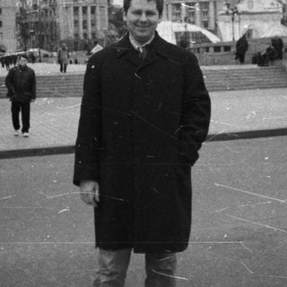 Man stands in a plaza with buildings and people.