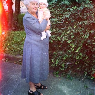 Woman in coat holds baby outdoors in a garden.
