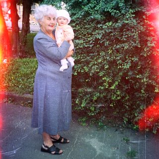 Woman in coat holds baby outdoors in a garden.