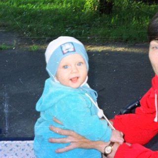 Woman holds baby outdoors, with greenery and buildings in background.