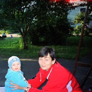 Woman holds baby outdoors, with greenery and buildings in background.