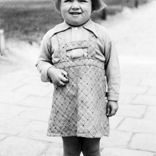 Young child stands outdoors in a patterned dress and socks.
