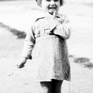 Here's a brief summary of the image: Young child in dress stands outside a building.