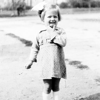 Here's a brief summary of the image: Young child in dress stands outside a building.