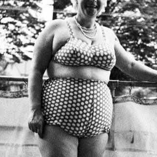 Woman in vintage swimsuit stands on a balcony.