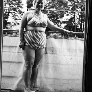 Woman in vintage swimsuit stands on a balcony.