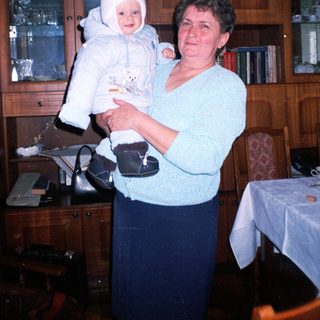 Woman holds baby indoors, standing near furniture and cabinets.