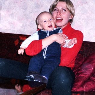Woman holds baby on a red couch in a home.