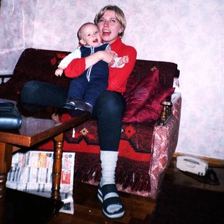 Woman holds baby on a red couch in a home.