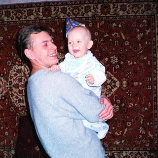 Man holds baby, both smiling, against a patterned rug.