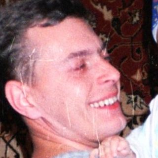 Man holds baby, both smiling, against a patterned rug.