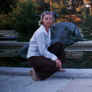 Woman kneels near a fountain with trees and building.