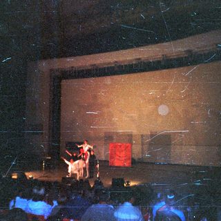 Concert stage with performers, audience visible in the darkness.