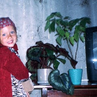 Child in red suit stands near plants, indoors.