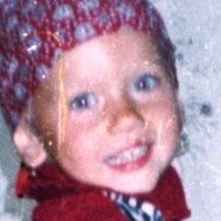 Child in red suit stands near plants, indoors.