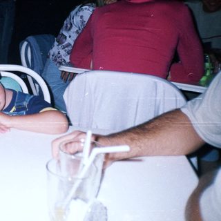 Boy sleeps on table with drinks and people nearby.