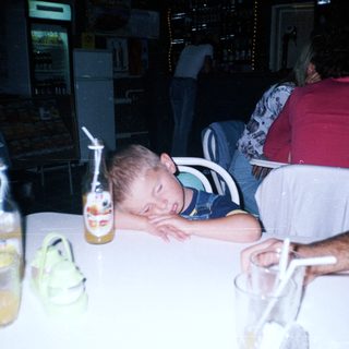 Boy sleeps on table with drinks and people nearby.