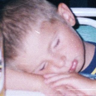 Boy sleeps on table with drinks and people nearby.