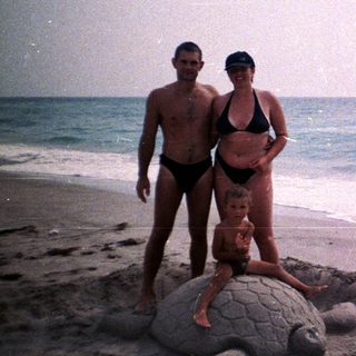 Family poses near a sand turtle sculpture on a beach.